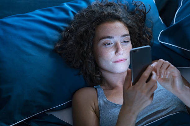 wanita muda menggunakan smartphone sambil berbaring di tempat tidur / iStockphoto